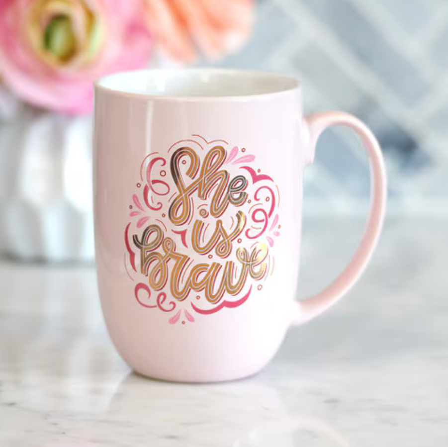 Pink ceramic coffee mug with gold foil lettering saying "She is Brave," featuring Christian Art Gifts branding and a stylish drinkware design.